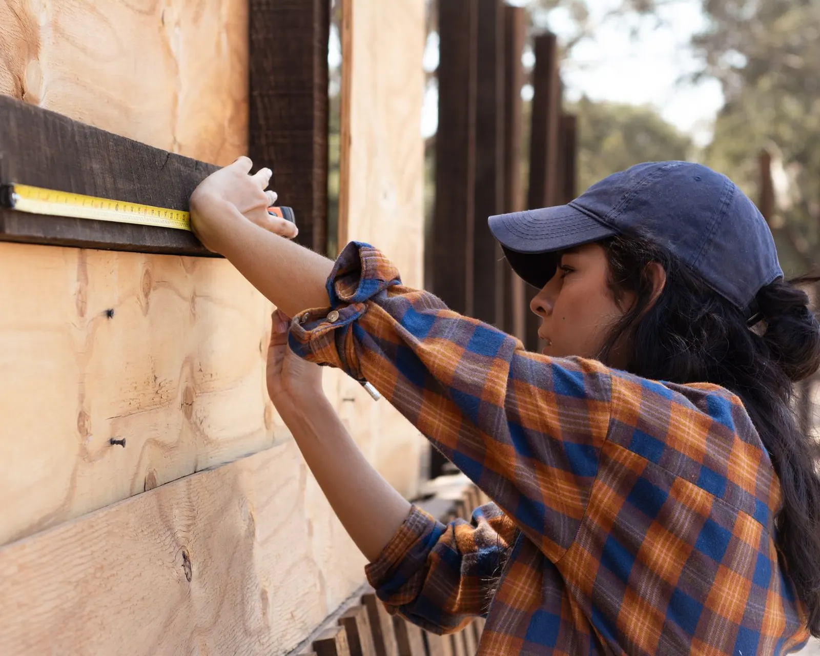 Carpenter in a cap measuring a timber member with a yellow tape measure on an outdoor frame
