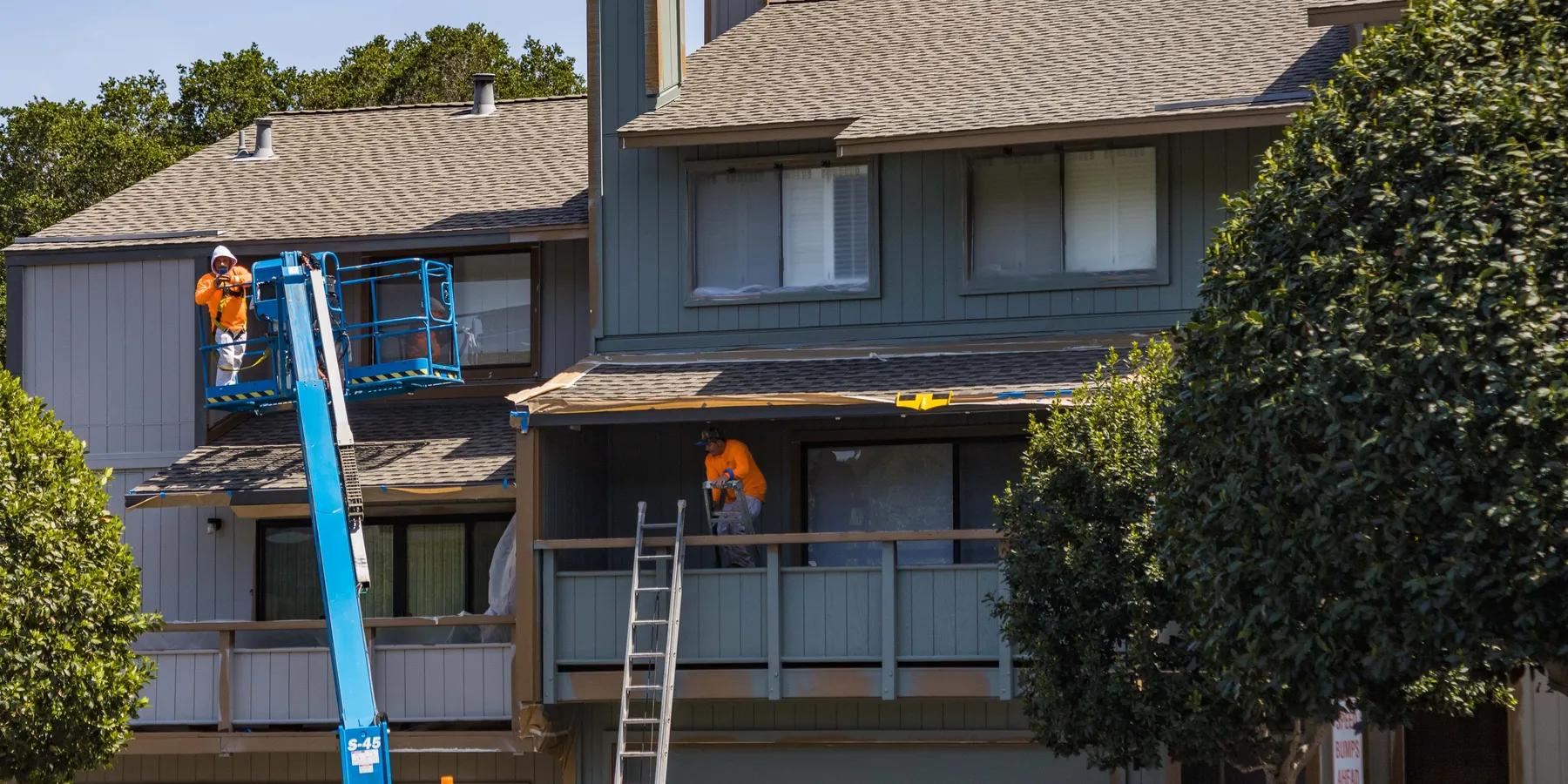 Painters on a boom lift and a balcony preparing the grey-green wood siding of a two-story house for painting