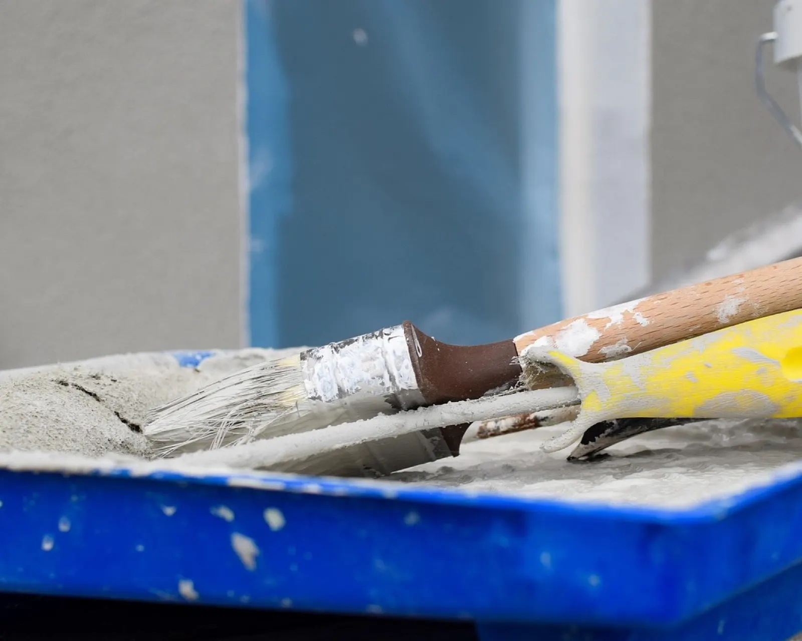 Paint brush and roller handle resting in a blue paint tray in front of a house facade being painted outdoors