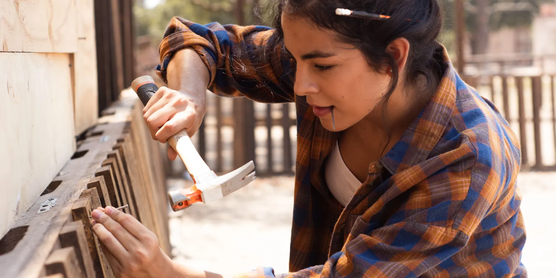 Carpenter in a plaid shirt hammering a nail into the boards of a wooden fence as it is built