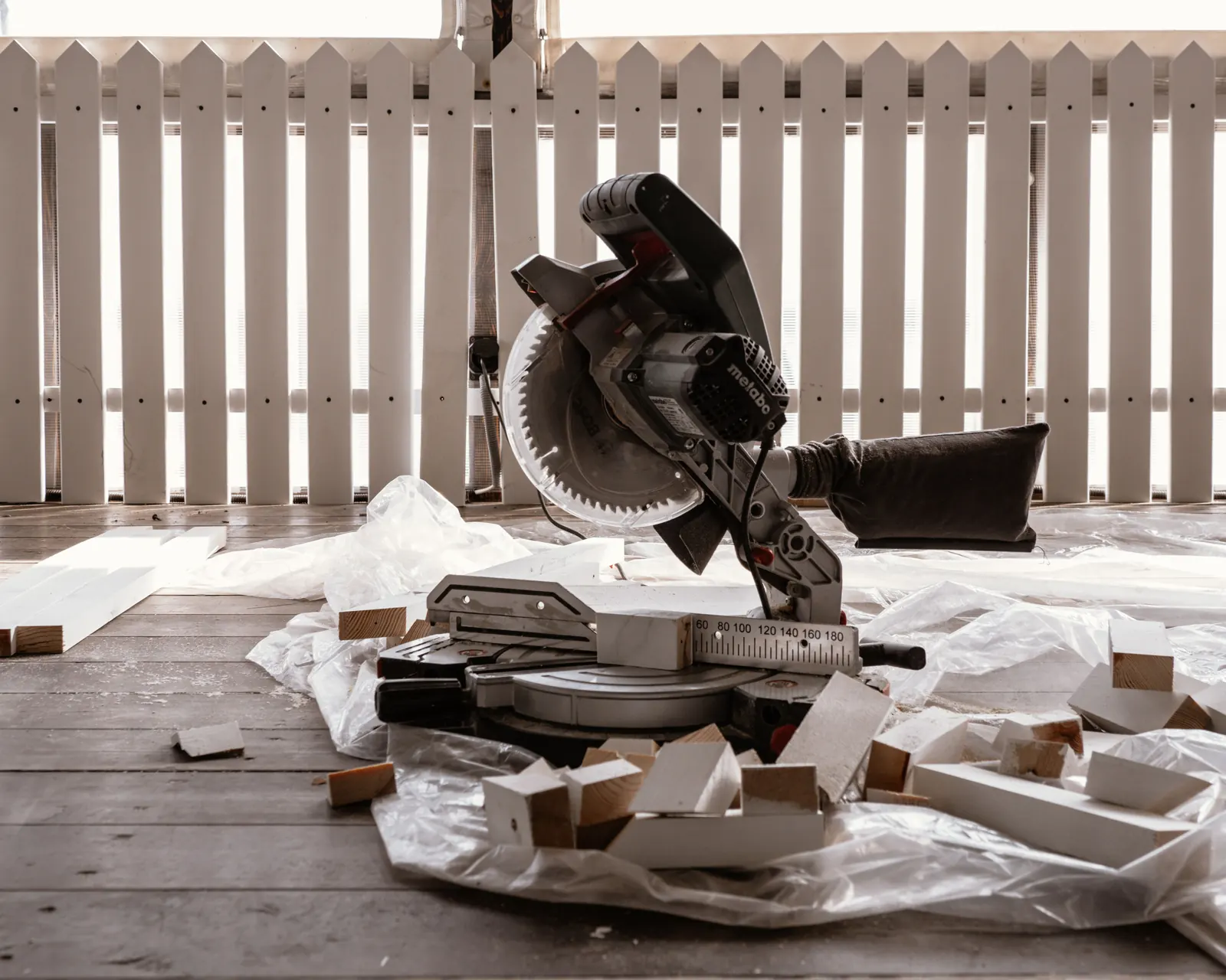 Mitre saw and offcuts of white-painted boards on a plastic sheet in front of a white picket fence, the cutting station for building a fence