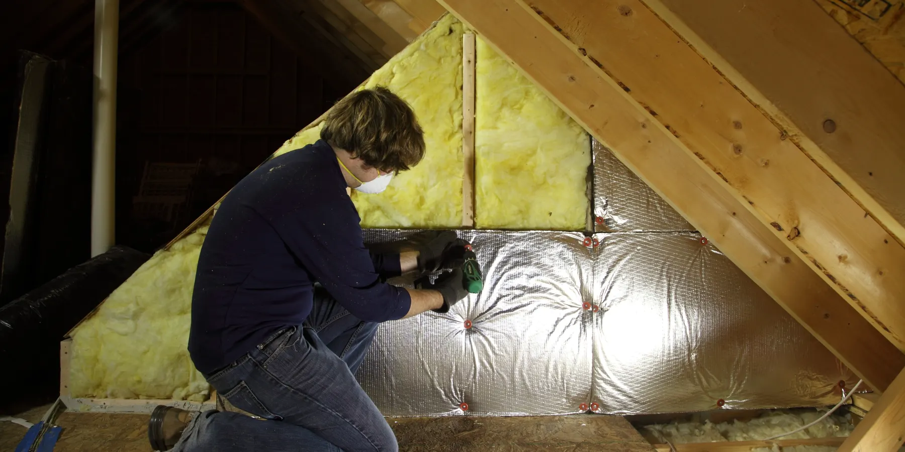 A homeowner in a dust mask fitting fiberglass batts and foil bubble insulation on an attic knee wall