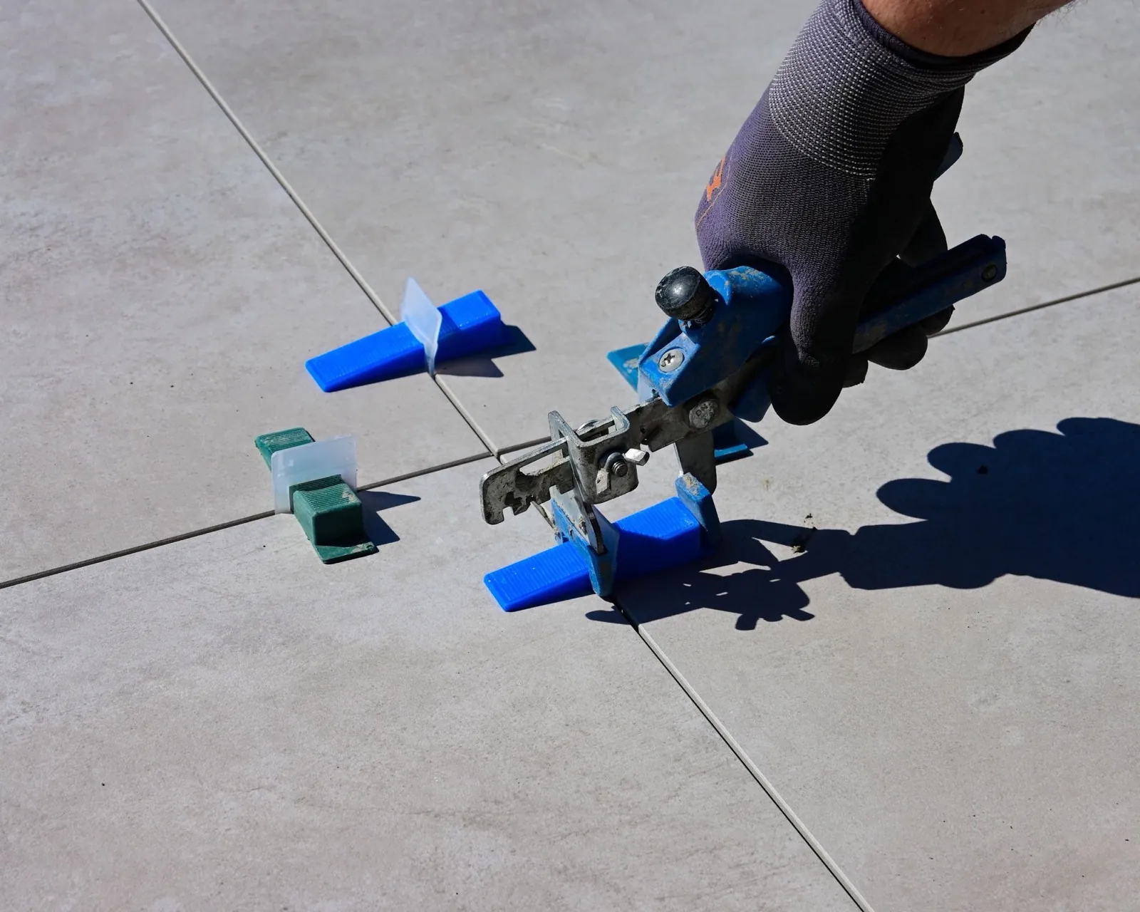 Gloved hand using a tile leveling tool on large gray tiles with blue and green leveling clips beside it